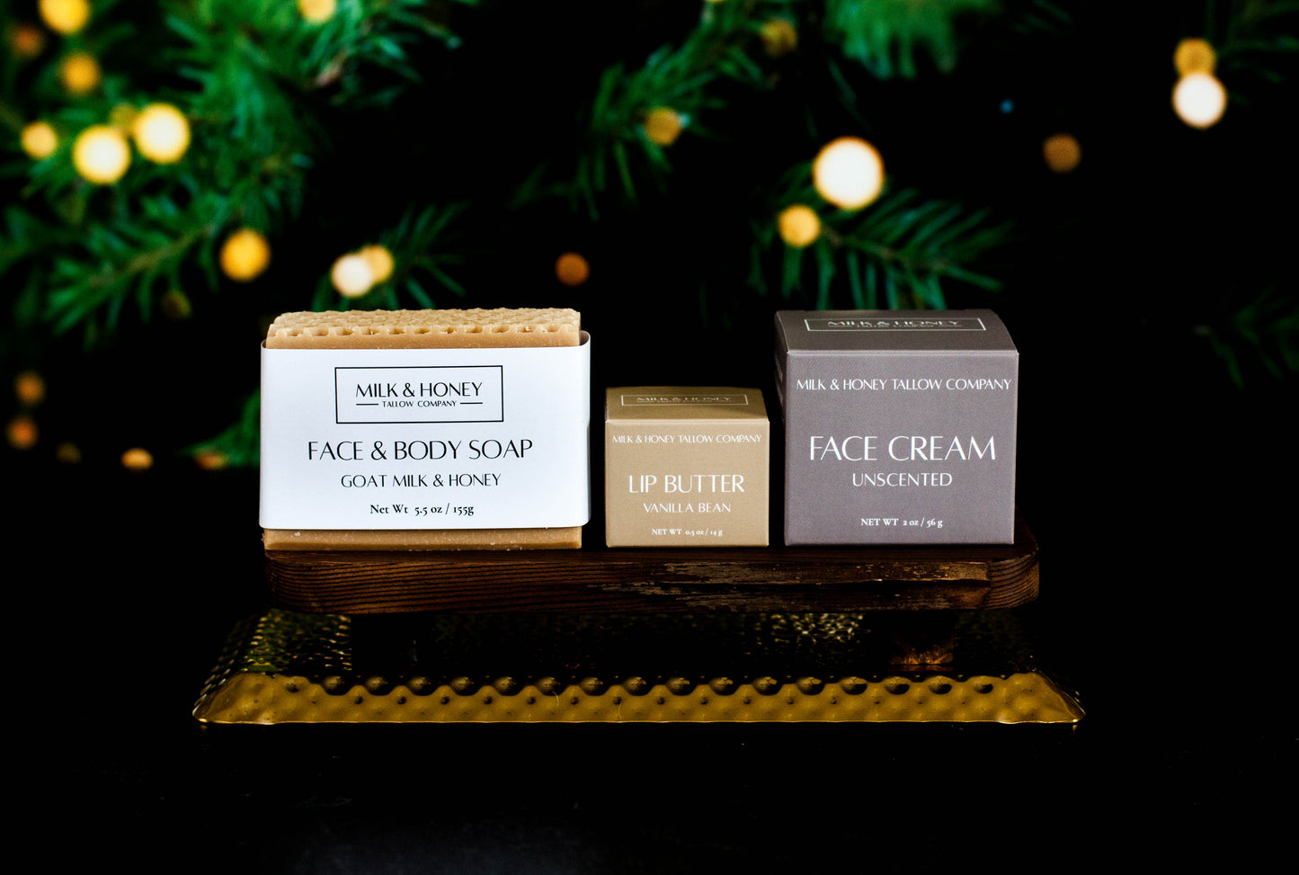 Set of natural tallow skincare products including soap, lip butter, and face cream on a decorative stand with blurred Christmas tree lights in the background.