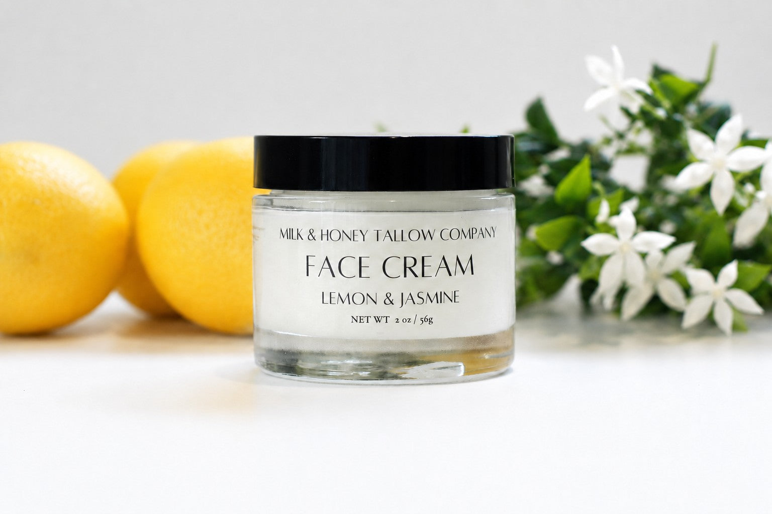 A jar of Milk & Honey Fallow Company Face Cream in Lemon & Jasmine scent, styled with fresh lemons and white flowers on a clean white background.