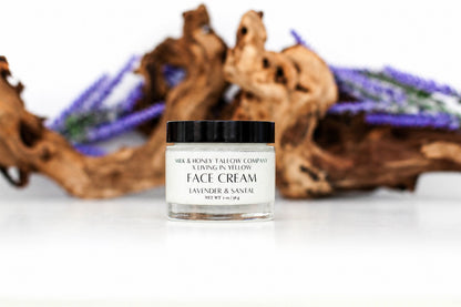 A small clear jar of face cream with a black lid and white label reading 'Milk & Honey Tallow Company A Living in Yellow Face Cream Lavender & Sandal' sits on a white surface with blurred brown driftwood and purple flowers in the background.
