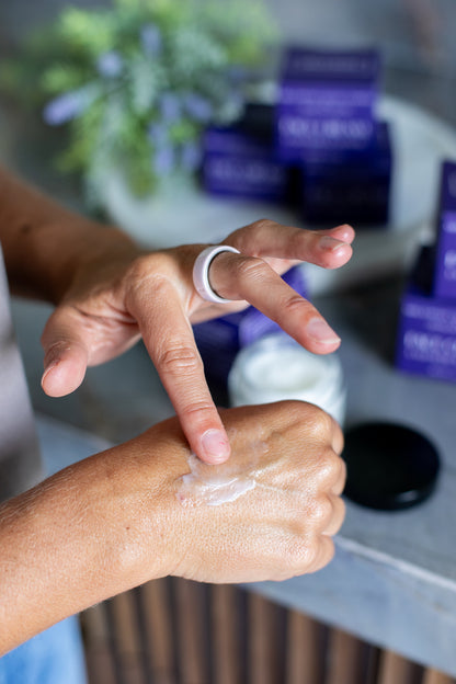 Person applying lavender & santal luxury face cream to their hand with a blurred background of skincare products.