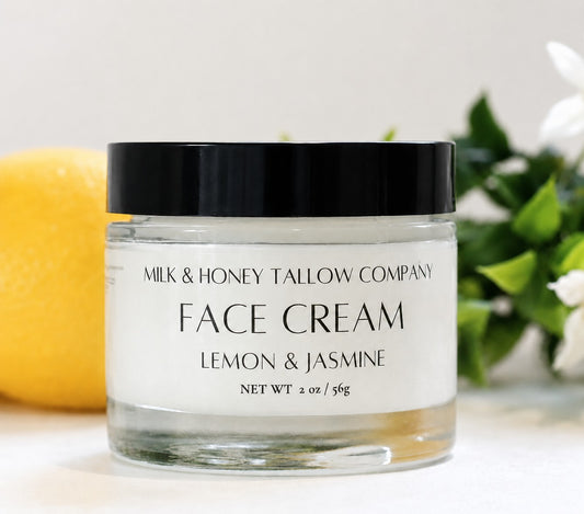 A clear glass jar of Milk & Honey Tallow Company Face Cream in the Lemon & Jasmine scent, featuring a black lid and white label, styled with a lemon and green leaves in the background.