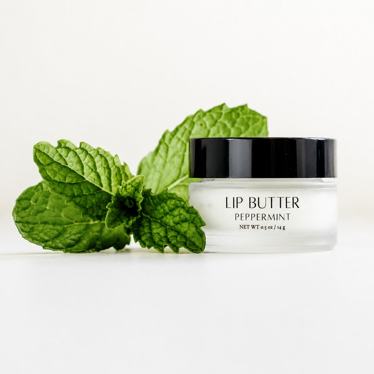A jar of lip butter labeled 'LIP BUTTER PEPPERMINT' with a black lid and clear container, styled with fresh green peppermint leaves on a clean white background.