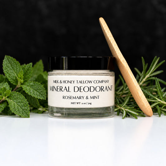 A jar of Milk & Honey Tallow Company Mineral Deodorant, Rosemary & Mint, styled with fresh rosemary and mint leaves and a bamboo applicator on a white surface against a dark background.