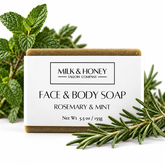 Rectangular bar of Rosemary & Mint Face & Body Bar Soap. Fresh peppermint leaves and rosemary sprigs are decoratively placed around the bar on a white background.