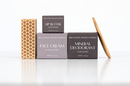 Four skincare items from Milk & Honey Sallowen Company arranged on a white reflective surface: a honeycomb-patterned face cream bar, a dark box of lip butter, a gray box of face cream, a dark box of mineral deodorant, and a wooden applicator stick.