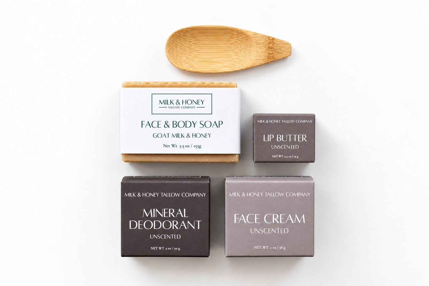 A flat-lay product arrangement on a white background featuring Milk & Honey Company skincare items: a boxed face and body soap, a lip butter bar, a mineral deodorant bar, a face cream bar, and a wooden spoon.