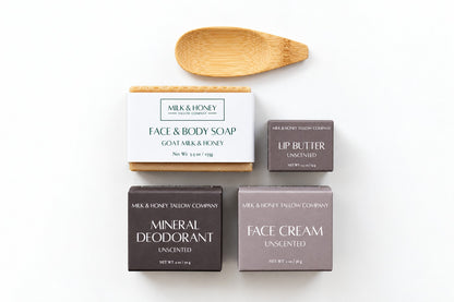 A flat-lay product arrangement on a white background featuring Milk & Honey Company skincare items: a boxed face and body soap, a lip butter bar, a mineral deodorant bar, a face cream bar, and a wooden spoon.