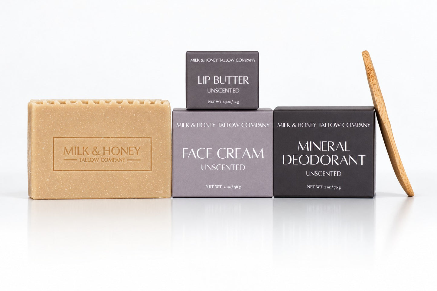 Four Milk & Honey Tallow Company skincare items arranged on a white reflective surface: a tan unscented soap bar, a dark gray unscented lip butter box, a medium gray unscented face cream box, a dark charcoal unscented mineral deodorant box, and a wooden stick tool leaning against the right side.