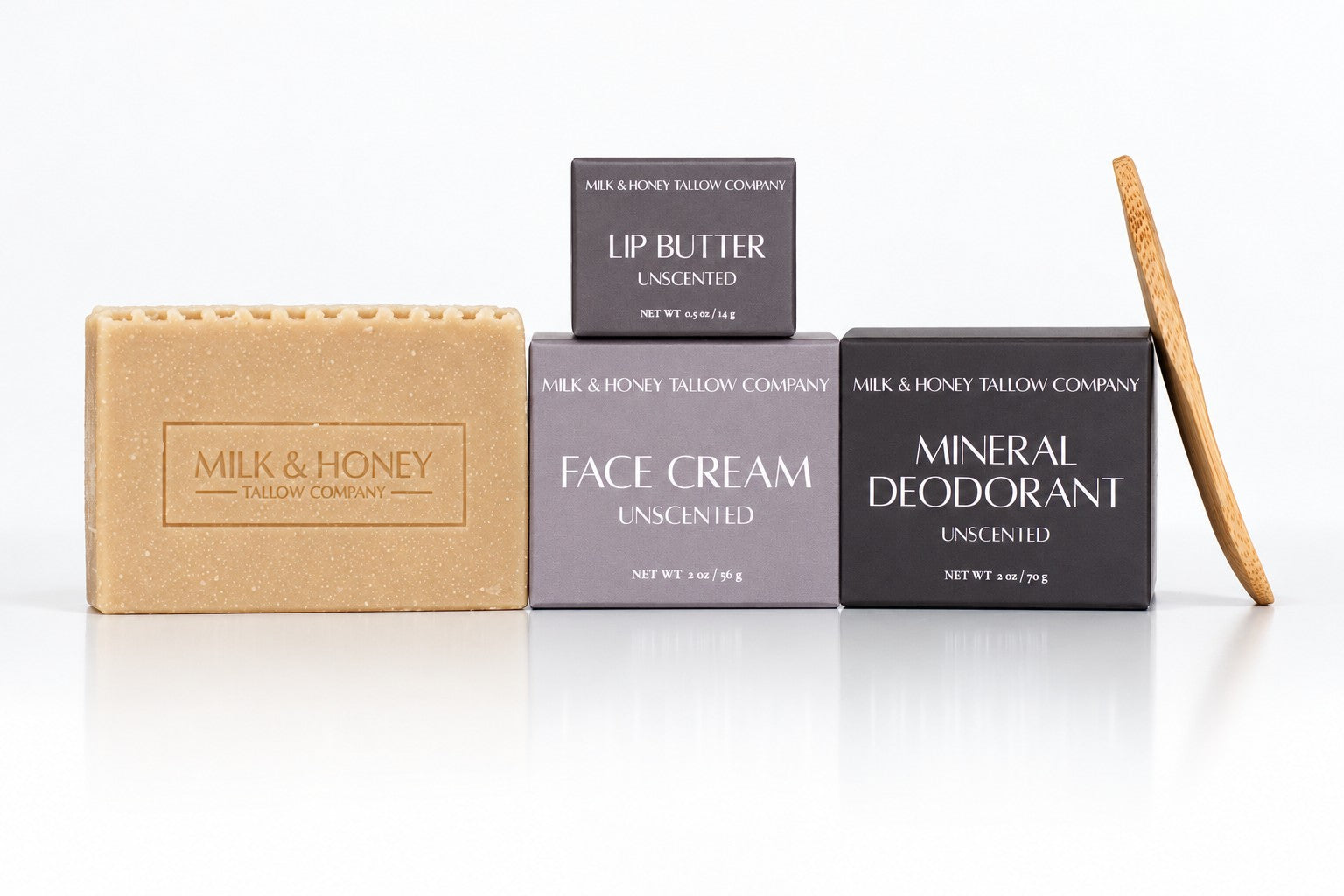 Four Milk & Honey Tallow Company skincare items arranged on a white reflective surface: a tan unscented soap bar, a dark gray unscented lip butter box, a medium gray unscented face cream box, a dark charcoal unscented mineral deodorant box, and a wooden stick tool leaning against the right side.