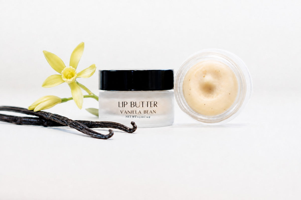 A jar of lip butter labeled 'LIP BUTTER VANILLA BEAN' with a black lid, shown next to an open jar of creamy luxury lip butter and vanilla beans with a yellow vanilla flower on a white background.